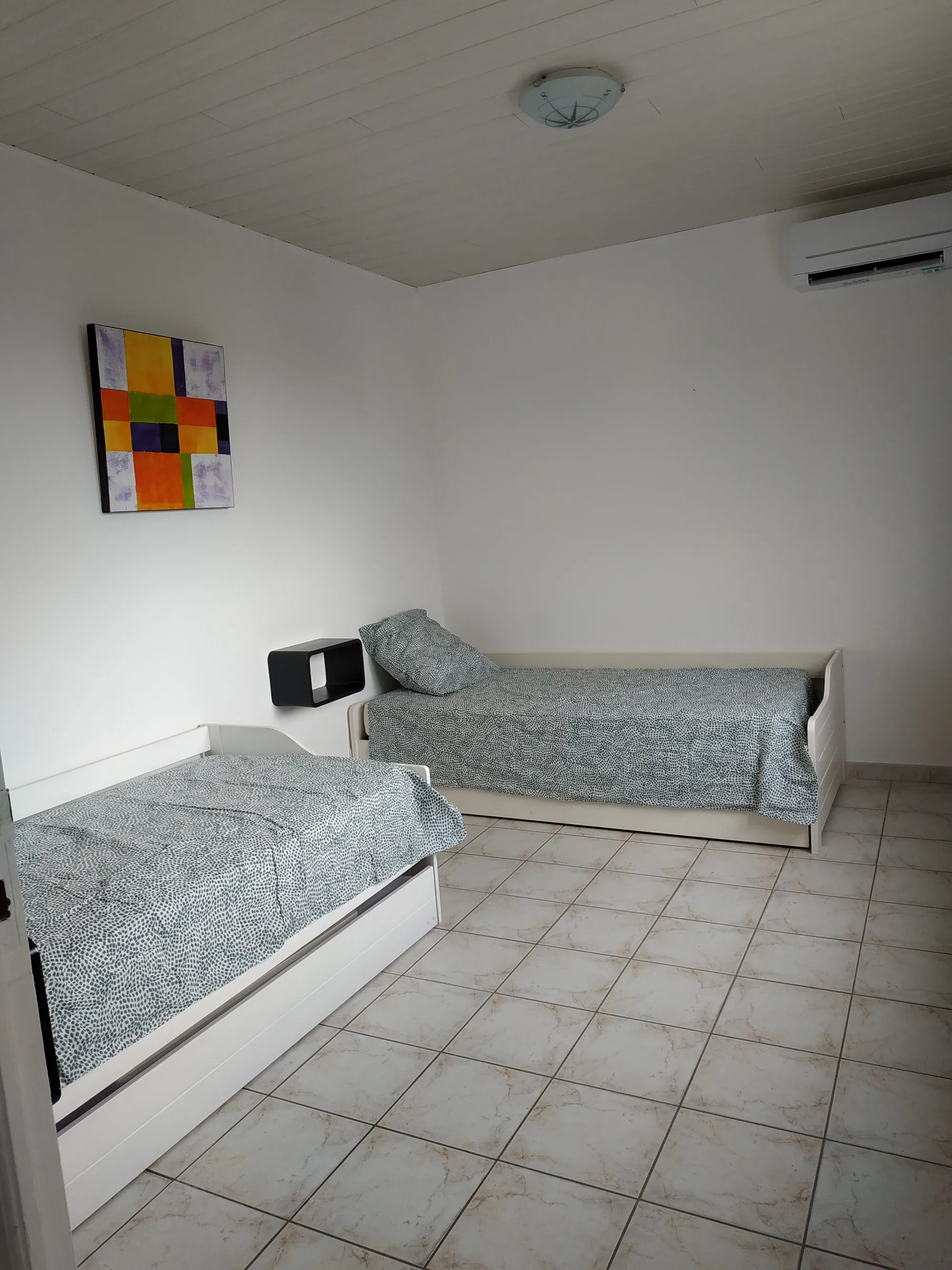 Deux lits simples dans une chambre claire, avec un tableau coloré au mur et un climatiseur