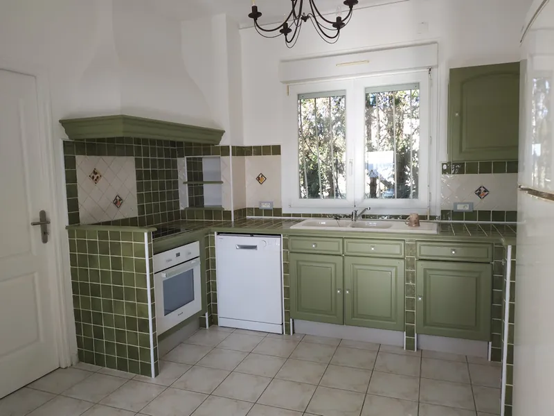 Cuisine équipée à carrelage et façades vert olive, avec four, lave-vaisselle et fenêtre sur le jardin