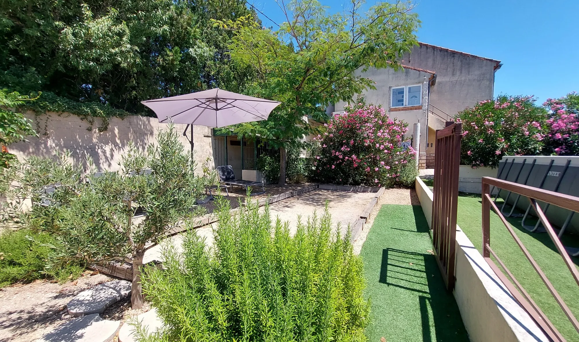 Le jardin avec un parasol, des lauriers-roses en fleur, un olivier et l'entrée du logement
