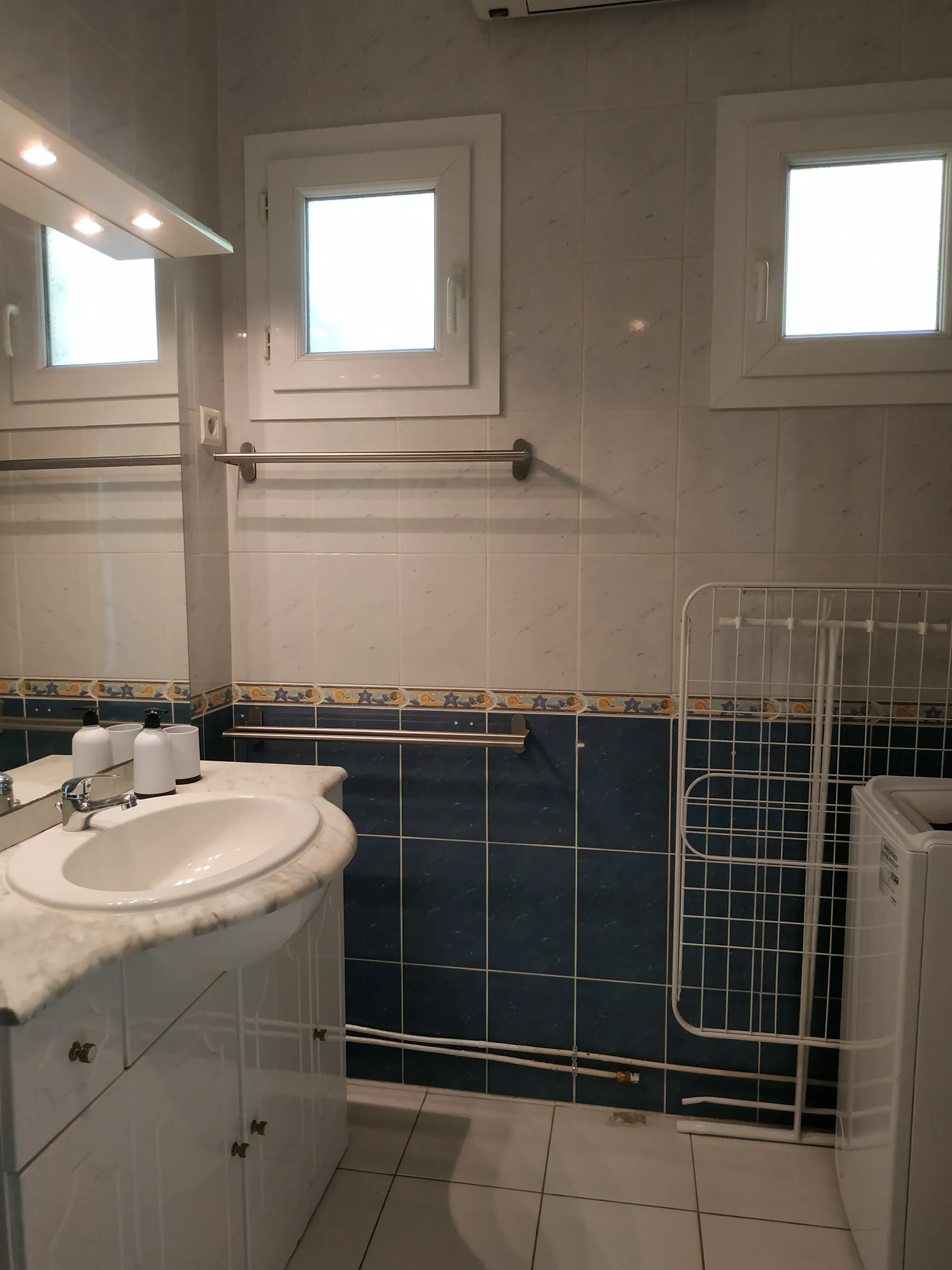 La salle de bains : vasque sur meuble blanc, carrelage bleu et deux petites fenêtres, avec un étendoir à droite