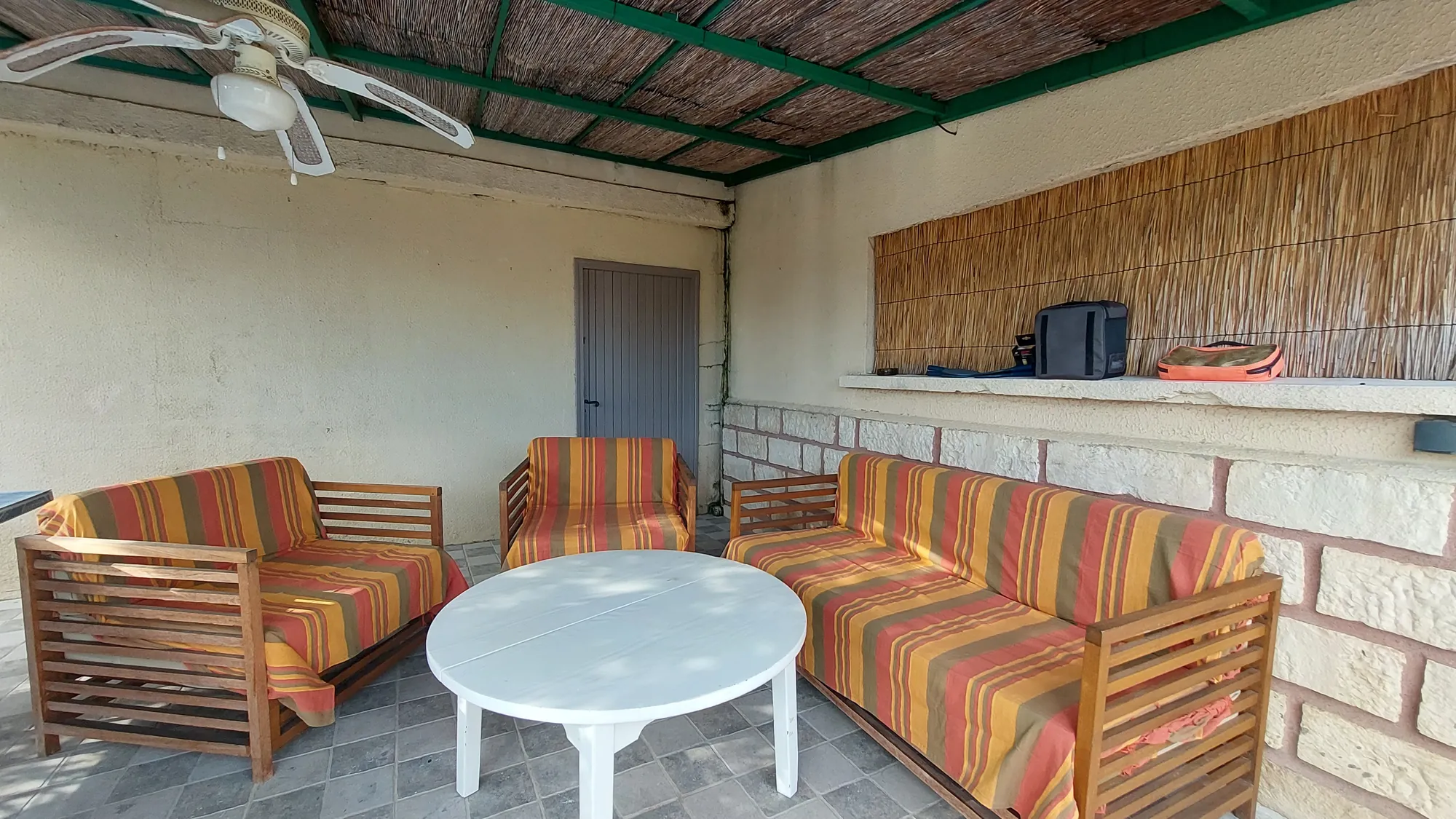Le salon de jardin du patio : deux canapés et un fauteuil à rayures autour d'une table ronde blanche, sous un ventilateur de plafond
