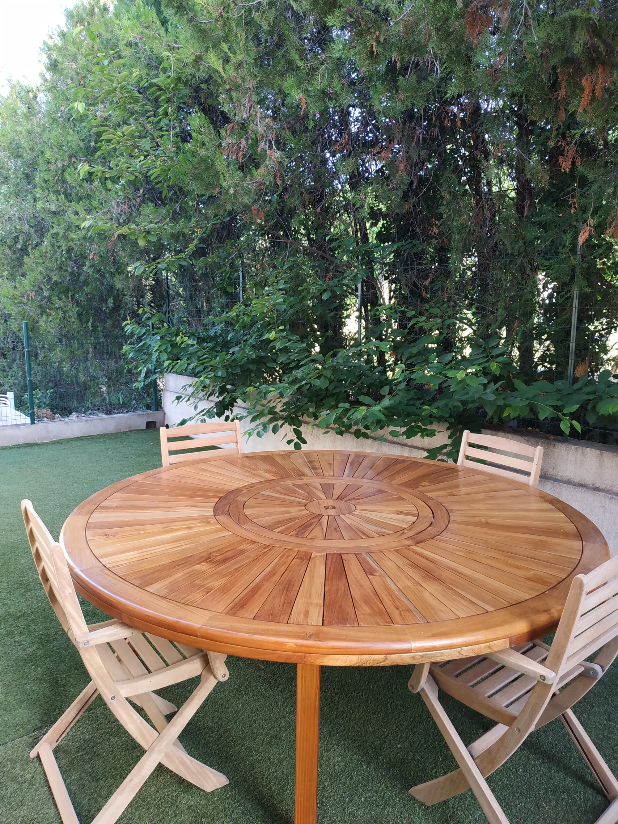 Table ronde en teck et quatre chaises pliantes, sur la pelouse devant une haie