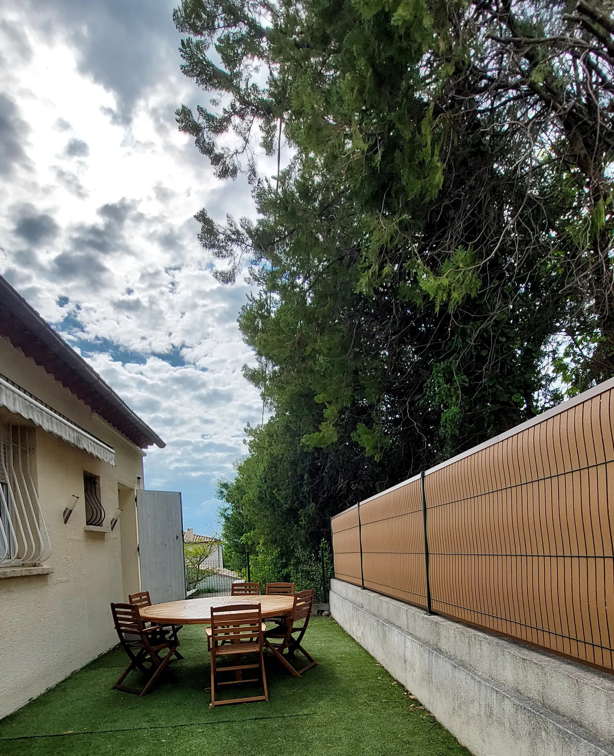 La table ronde en teck et ses chaises sur la pelouse, le long du muret et de la clôture, à côté de la maison